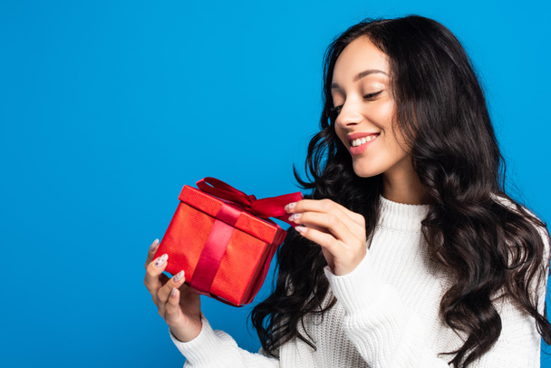happy woman in sweater looking at christmas present isolated on blue - Photo, image