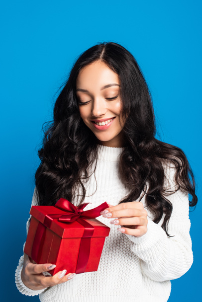 happy woman in sweater touching ribbon on christmas present isolated on blue - Фото, зображення