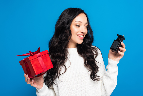 femme heureuse en pull tenant boîte cadeau de Noël et regardant bouteille avec parfum isolé sur bleu - Photo, image