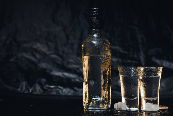 Close-up view of bottle of vodka with glasses standing on ice on black - Photo, image