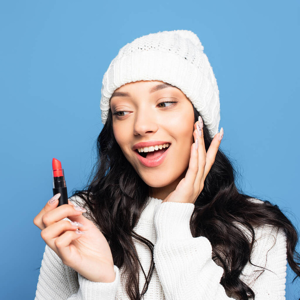 happy brunette woman in hat looking at lipstick isolated on blue - Photo, Image