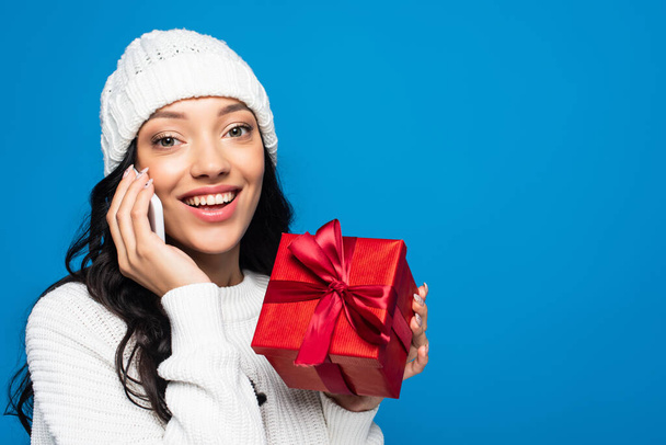 happy woman in knitted hat holding wrapped present and talking on smartphone isolated on blue - Фото, изображение