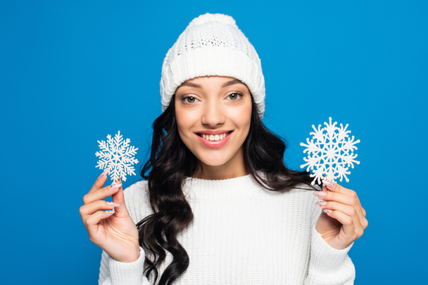 happy woman in knitted hat holding decorative snowflakes isolated on blue - Fotó, kép