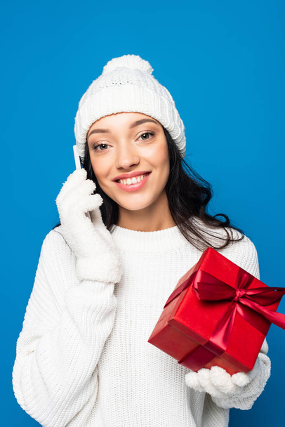 pleased woman in knitted hat and gloves holding gift box and talking on smartphone isolated on blue - Foto, Imagem