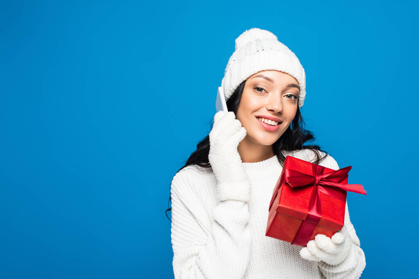 happy woman in knitted hat and gloves holding gift box and talking on smartphone isolated on blue - Photo, Image