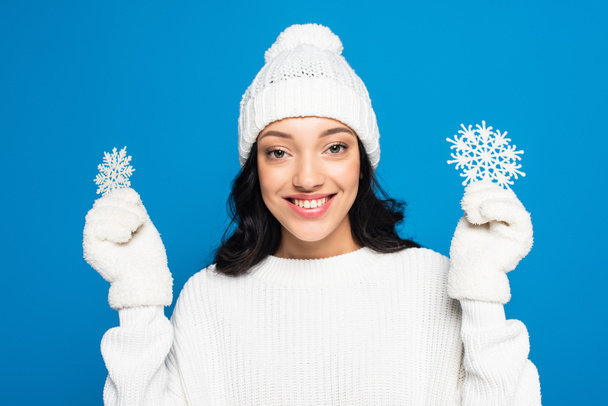 boldog nő kötött kalapban és kesztyűben kezében dekoratív hópelyhek elszigetelt kék - Fotó, kép