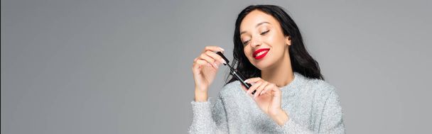 happy brunette woman with red lips looking at mascara isolated on grey, banner - Photo, Image