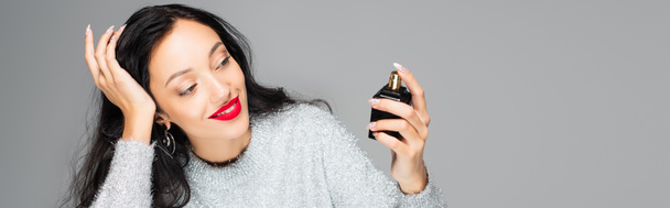 pleased woman with red lips holding bottle with perfume isolated on grey, banner - Фото, изображение
