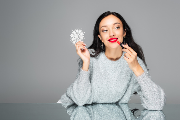 happy woman with red lips holding decorative snowflake and applying lipstick isolated on grey - Photo, Image