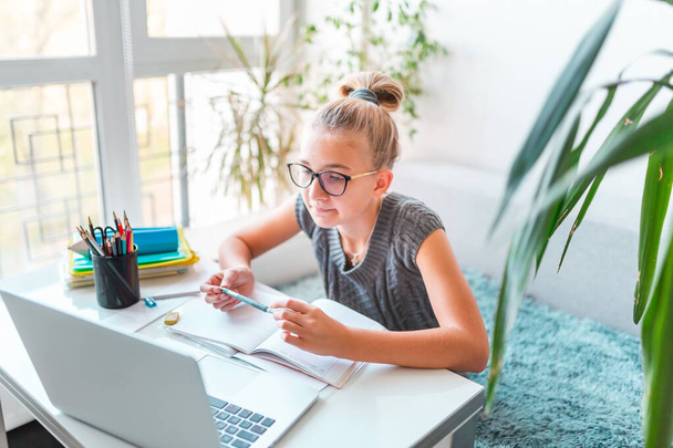 Beautiful young school girl left-handed working at home in her room with laptop and class notes studying in a virtual class. Distance and online education, e-learning during quarantine - 写真・画像