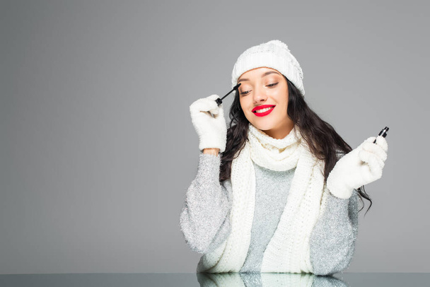 радісна і брюнетка жінка в зимовому вбранні наносить туш ізольовано на сірий
 - Фото, зображення