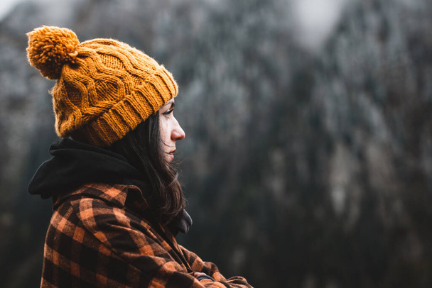 Vista lateral de la joven reflexiva con camisa a cuadros y sombrero de invierno amarillo mirando a la vista en condiciones de clima frío. Viajes, aire libre y concepto de clima frío. Copiar espacio - Foto, imagen