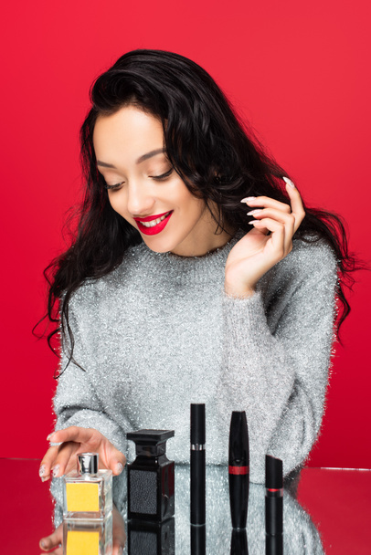 usmívající se žena ve svetru při pohledu na dekorativní kosmetiku a láhve s parfémem izolované na červené - Fotografie, Obrázek