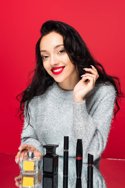 mujer complacida en suéter mirando a la cámara cerca de cosméticos decorativos y botellas con perfume aislado en rojo - Foto, imagen