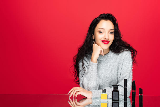 young pleased woman in sweater looking at camera near decorative cosmetics and bottles with perfume isolated on red - Foto, afbeelding