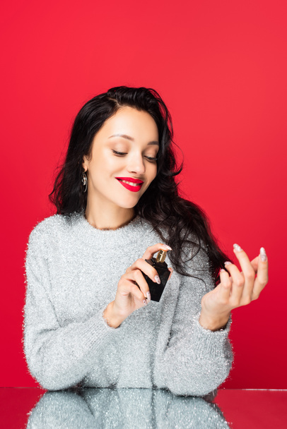 pleased woman holding bottle and applying perfume isolated on red - Foto, Imagem