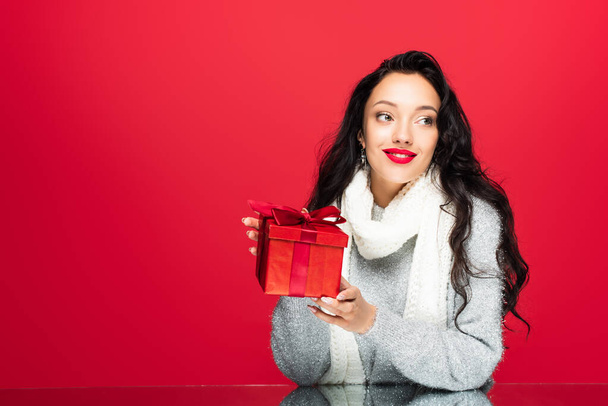 joyful and brunette woman in sweater holding christmas present isolated on red  - Fotó, kép