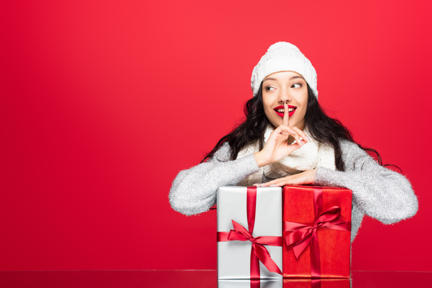 joyful woman in hat and warm scarf showing hush sign near presents isolated on red - Photo, Image