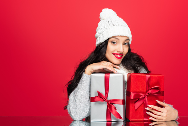 mujer feliz en sombrero abrazando regalos de Navidad aislados en rojo - Foto, imagen
