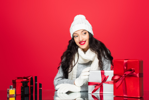 mujer alegre en sombrero, guantes y bufanda caliente cerca de regalos de Navidad y cosméticos decorativos aislados en rojo  - Foto, Imagen