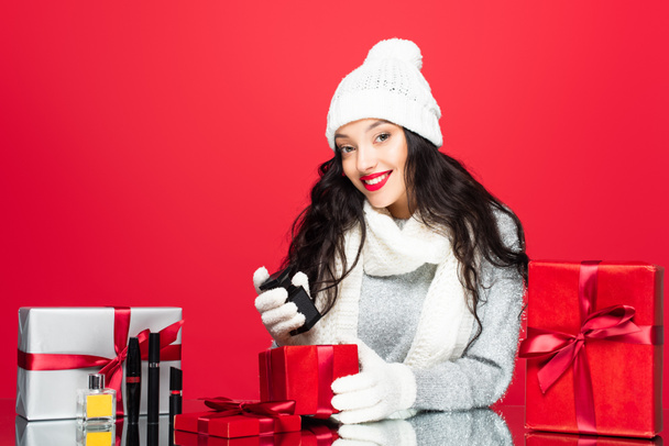 fröhliche Frau mit Mütze, Handschuhen und warmem Schal, die eine Flasche mit Parfüm in der Nähe von Weihnachtsgeschenken und dekorativer Kosmetik auf Rot hält  - Foto, Bild