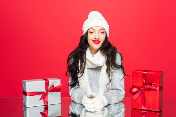 joyful woman in winter outfit smiling near christmas presents isolated on red - 写真・画像
