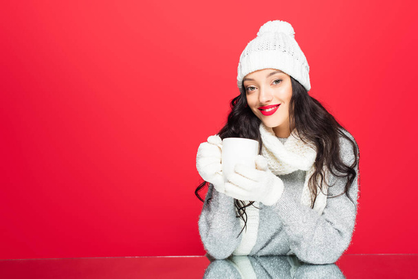 happy woman in winter outfit holding cup with beverage isolated on red - Foto, afbeelding