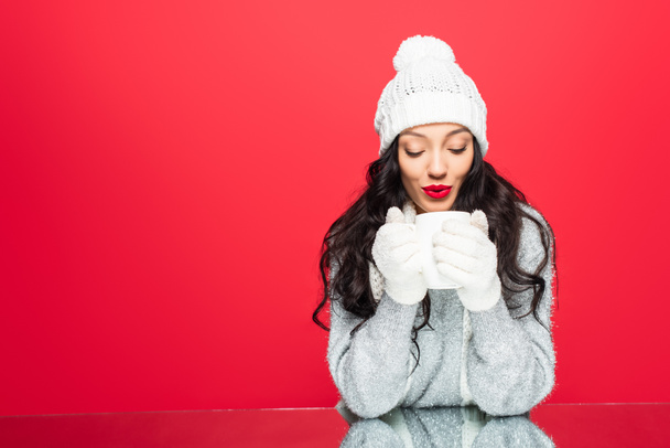 pleased woman in winter outfit blowing on cup with beverage isolated on red - Fotografie, Obrázek