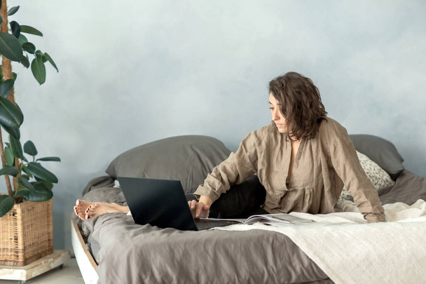 Une jeune femme brune utilise un ordinateur portable dans sa chambre alors qu'elle est assise sur le canapé. Utilise un ordinateur portable pour regarder un film, discuter avec des amis, lire un livre et voir un e-mail. - Photo, image