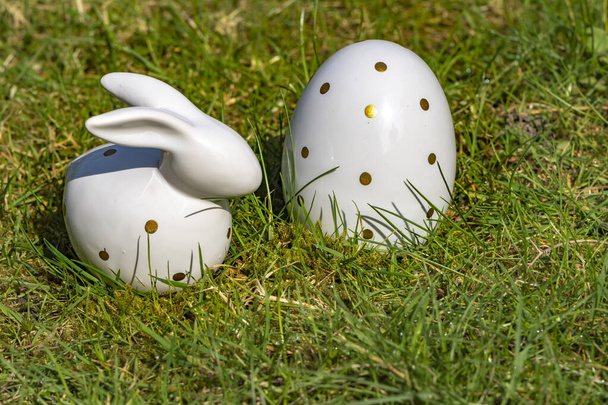 Húsvéti nyuszi és húsvéti tojás fehér kerámiából arany polka pontokkal a réten. - Fotó, kép