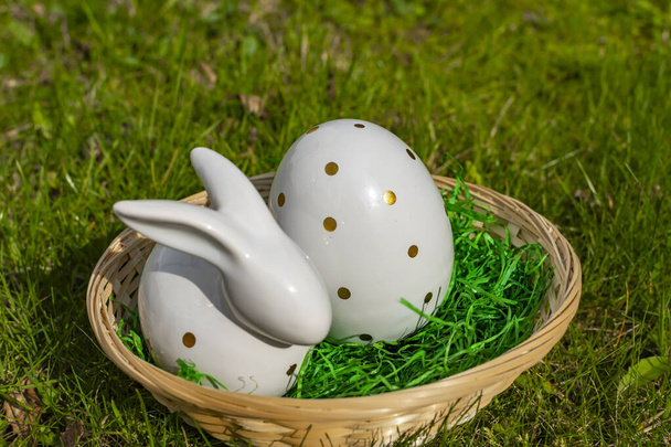 Um coelho de Páscoa e um ovo de Páscoa feito de cerâmica branca com bolinhas douradas em uma cesta em um prado. - Foto, Imagem