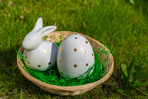 Egy húsvéti nyuszi és egy húsvéti tojás fehér kerámiából arany pöttyökkel egy kosárban egy réten.. - Fotó, kép