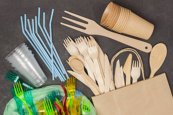Wooden forks and knives in paper bag. Colored plastic forks, glasses, beverage straws in plastic bag. Plastic and eco concept.  Ecology environmental care. Black background. Flat lay. - Foto, Imagem