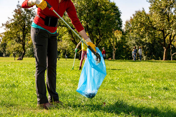 People who collect plastic and paper in the park. Volunteer woman with yellow gloves and grabber picking up garbage for cleaning at the park. Ecology concept - Photo, image