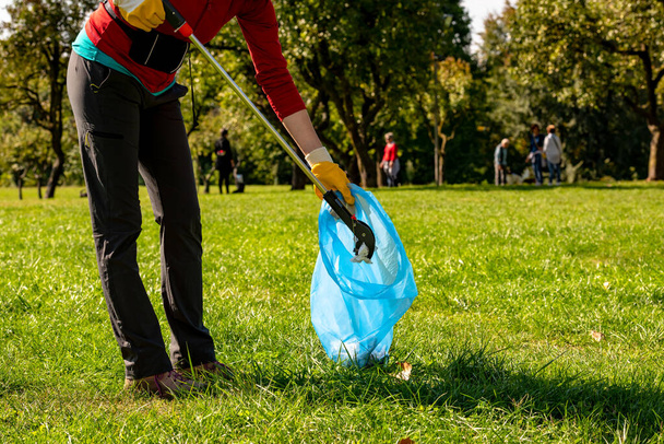 People who collect plastic and paper in the park. Volunteer woman with yellow gloves and grabber picking up garbage for cleaning at the park. Ecology concept - Foto, Imagem