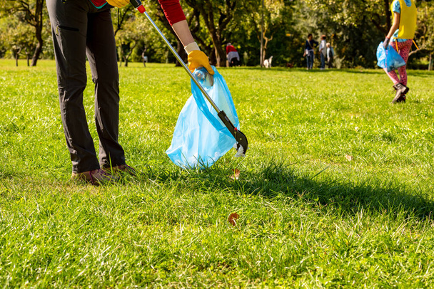 People who collect plastic and paper in the park. Volunteer woman with yellow gloves and grabber picking up garbage for cleaning at the park. Ecology concept - Foto, afbeelding