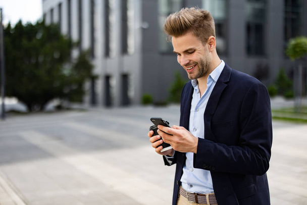 Handsome office worker holding smartphone and smiling. Happy young man using mobile phone apps, texting message, browsing internet, looking at smartphone. Coffee break near the business center - Foto, afbeelding
