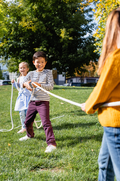 Smiling asian boy pulling rope while playing tug of war with friends on blurred foreground in park  - Photo, Image