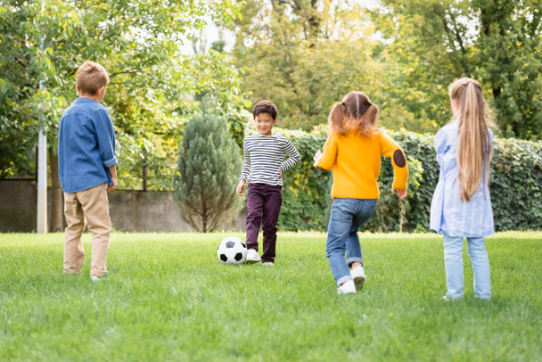 Cheerful asian boy playing football with friends on grassy lawn  - Photo, Image