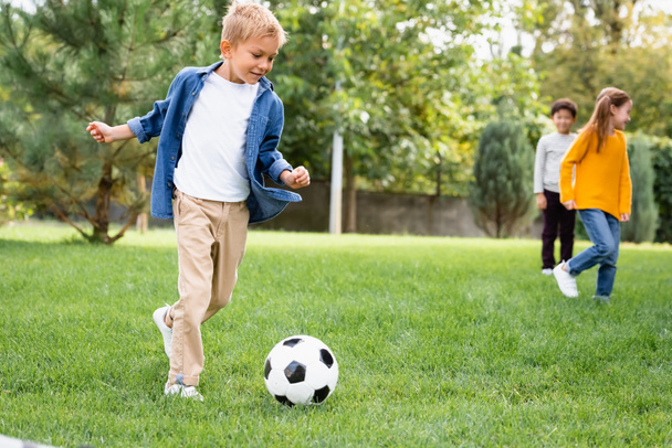 Smiling boy playing football near friends on blurred background in park - Foto, Imagen