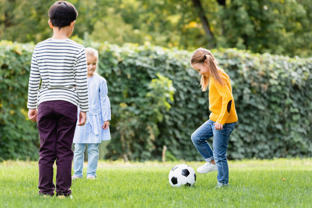 Улыбающаяся девушка играет в футбол рядом с друзьями на лужайке  - Фото, изображение