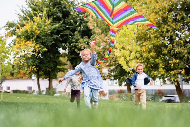 Smiling girl holding flying kite while playing with boys on blurred background in park  - Фото, зображення
