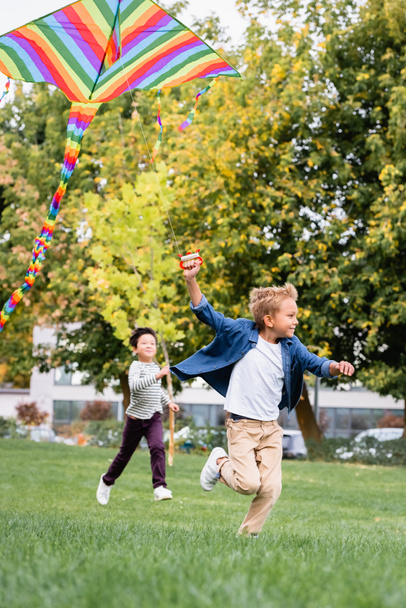 sonriente chico holding volando cometa mientras corriendo cerca asiático amigo en parque  - Foto, Imagen