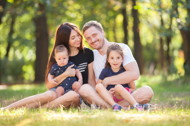 Happy mother, father, son and daughter in the park. Beauty nature scene with family outdoor lifestyle. Happy family resting together sitting on the grass, having fun outdoor. Happiness and harmony in family life - 写真・画像