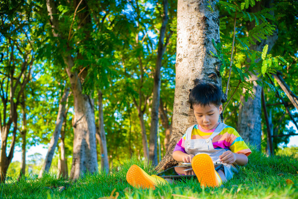 Piccolo ragazzo asiatico seduto su erba verde sotto albero uso tablet computer internet browser in città parco luce del tramonto - Foto, immagini
