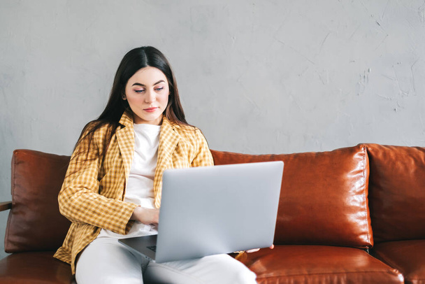 Portrait of focused beautiful woman sitting on a sofa, looking at laptop screen on video call or chat and typing on keyboard. Businesswoman working distantly at home office. - Photo, Image