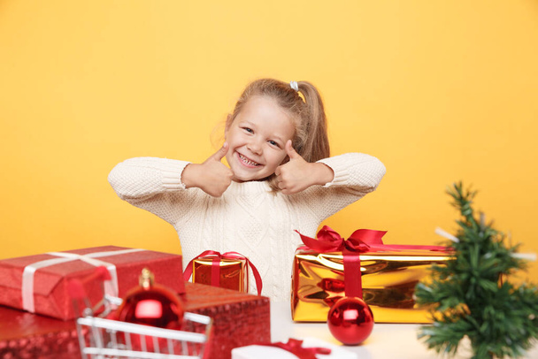 Gelukkig meisje in trui zit met geschenken geïsoleerd boven de gele studio. Xmas-concept. - Foto, afbeelding