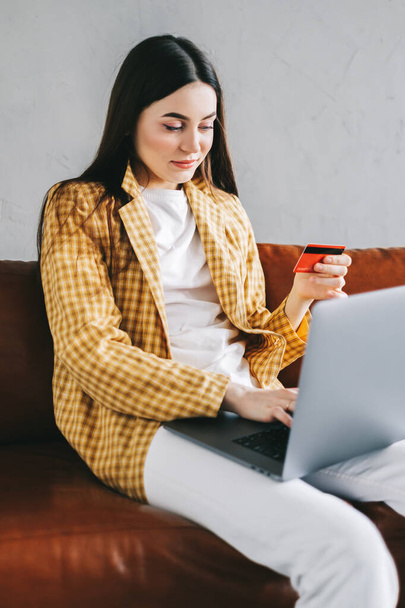 Young caucasian woman holding credit card, using laptop computer and shopping online. E-commerce, internet banking, spending money. High quality photo - Фото, изображение