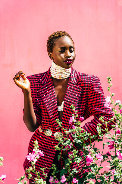 Hermosa modelo africana con piel negra sobre fondo rosa. Una sonrisa auténtica. Colores vibrantes, collar de caramelos. Foto de alta calidad. Flores rosadas. - Foto, imagen