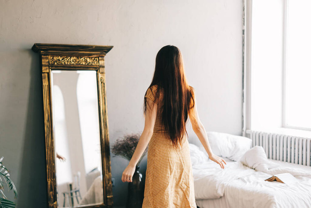Beautiful brunette woman in dress standing near big mirror in bedroom and looking on himself, fitting new outfit. High quality photo - 写真・画像